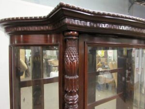 Close-up of a Spanish Revival curio cabinet showing the carved top molding, Greek key trim, and a central carved column beside the glass doors.