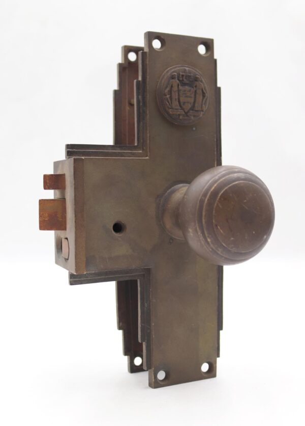 Art Deco bronze door knob set with a round knob and rectangular lock plate, shown in a close-up side view with mounting holes and visible surface wear from prior use.