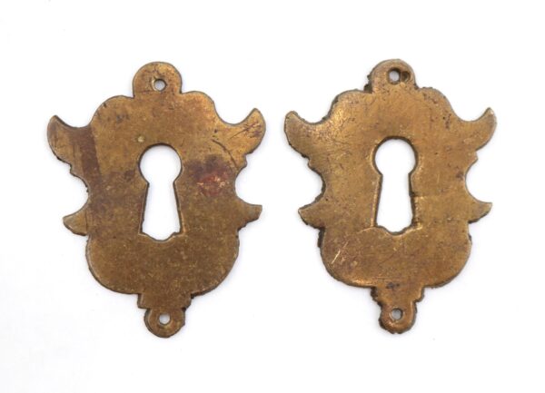 Pair of antique brass keyhole covers with a symmetrical flourish shape shown side by side, each with a central keyhole opening and top and bottom mounting holes, displaying heavy surface wear.