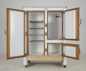 An open white icebox refrigerator with four doors, wood trim, wire shelves, and brass latches, shown with its interior compartments and surface wear visible.