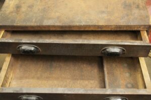 Close-up of a dark wood tone workshop chest of drawers with black steel bin pulls, showing open drawers and heavy surface wear.