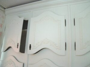 White built-in cabinet bank with curved doors, carved floral motifs, and dark original hardware, shown in a close-up of the upper and middle sections.