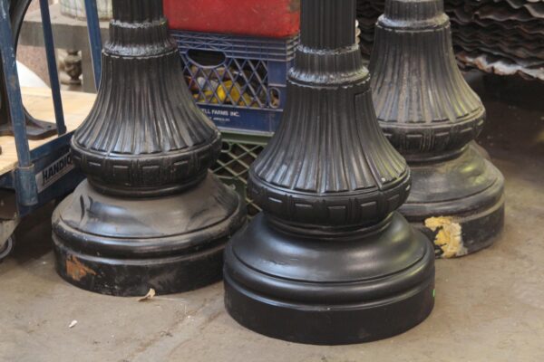 Three black street light post bases shown together in a close-up, with fluted cast aluminum detailing and rounded plinths, one displaying noticeable surface wear.