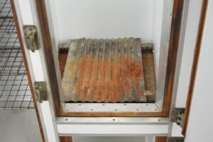 Open icebox refrigerator compartment with a corrugated metal shelf showing surface wear and rust discoloration, framed by white enamel panels and brass latch hardware.