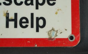Close-up of a steel safety wall sign with a red border, white field, and black text, showing the word “Help” and a predrilled mounting hole at the corner, with visible wear and stains from prior use.