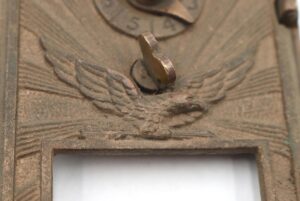 Close-up of a vintage bronze post office box door with a flying eagle emblem, combination dial, and open lower window cutout.