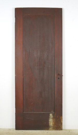 Vintage dark wood passage door with one full-length panel and an antique brass mortise lock, shown upright with visible surface wear and chips.