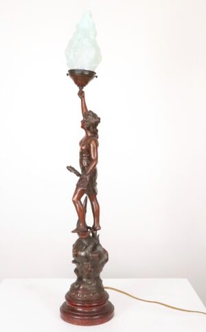 Art Nouveau table lamp with a dark wood-tone female figure holding a torch and standing on a crescent moon, topped with a white flame-shaped glass shade.