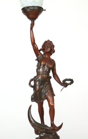 Art Nouveau lamp with a dark wood-tone female figure balancing on a crescent moon and holding a torch beneath an opaline flame glass shade.