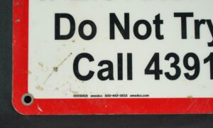 Close-up of a steel safety wall sign with a red border, white face, black text, and a predrilled mounting hole, showing surface wear and part of the warning message.