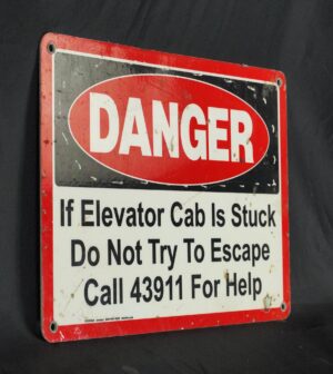 Steel safety wall sign with a red and black danger notice, four corner mounting holes, and text instructing, “If elevator cab is stuck, do not try to escape, call 43911 for help.”