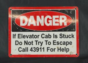 Industrial safety wall sign with a red and black danger notice, four mounting holes, and visible surface wear.