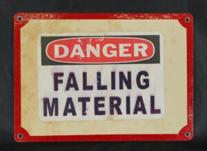 Industrial steel wall sign with a red border and four mounting holes, showing the warning text “Danger Falling Material” in black and red with visible surface wear.