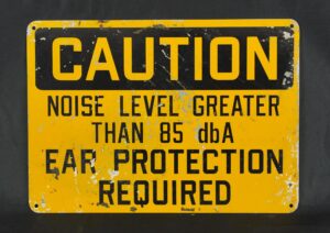 Industrial black and yellow steel caution sign with four mounting holes, reading “Noise level greater than 85 dBA, ear protection required,” shown with visible surface wear and chipped finish.
