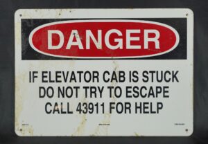 Rectangular steel safety sign with a red DANGER header, black warning text about a stuck elevator cab, four mounting holes, and visible surface wear and stains.