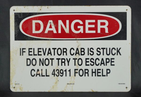 Rectangular steel safety sign with a red DANGER header, black warning text about a stuck elevator cab, four mounting holes, and visible surface wear and stains.