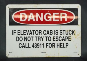 Industrial steel danger sign in black, red, and white with predrilled corner holes, reading “If elevator cab is stuck do not try to escape, call 43911 for help,” shown with surface wear and stains.