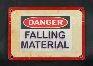 Industrial steel wall sign with a vinyl surface, red and white border, and four mounting holes, reading “Danger Falling Material.”