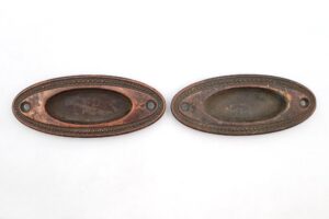 Pair of brass oval recessed window sash lifts shown side by side, each with a central pull recess and screw holes at both ends, with visible surface wear and rust.