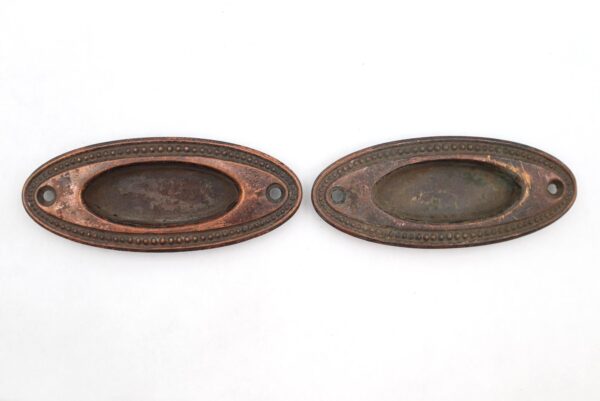 Pair of brass oval recessed window sash lifts shown side by side, each with a central pull recess and screw holes at both ends, with visible surface wear and rust.