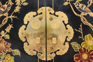 Close-up of a black lacquer cabinet door with hand-painted birds and flowers, polished brass hardware, and a central decorative medallion with round pulls.