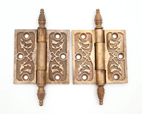 Pair of antique Victorian brass butt door hinges shown side by side, each with steeple tips, three knuckles, and an ornate template hole pattern with visible surface wear.