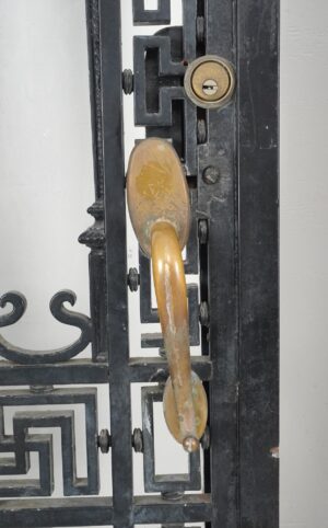Close-up of a wrought iron door showing an antique brass pull and lock, with Greek key detailing and surface wear visible on the black frame.