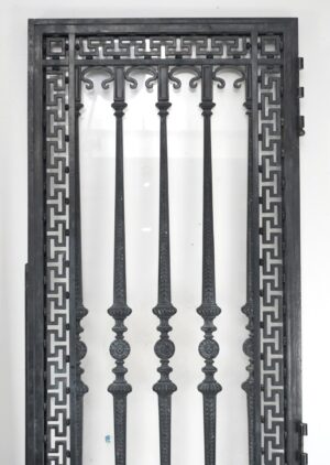Wrought iron entry door with a Greek key frame, vertical bars, and an ornate relief panel, shown in a front view with the right-side hinges visible.