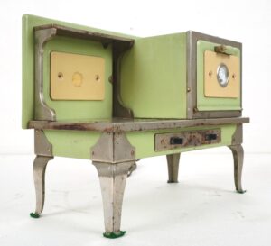 1930s toy stove in pale green and cream enamel with nickel-plated legs, shown at an angle with two front doors, one oven gauge, and worn paint.