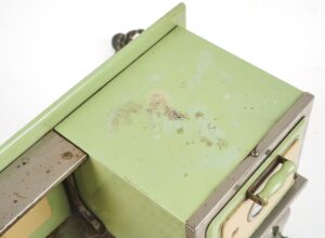 Close-up of a vintage toy stove in pale green and tan enamel with chipped paint and worn nickel trim.