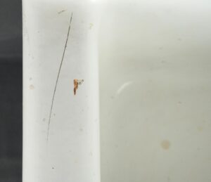 Close-up of a white fireclay sink basin showing surface wear, a long hairline crack, and a small rust chip in the porcelain glaze.