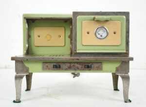 Vintage toy stove with two oven openings, cream panels, a round temperature dial, and worn green enamel on nickel-plated cabriole legs.
