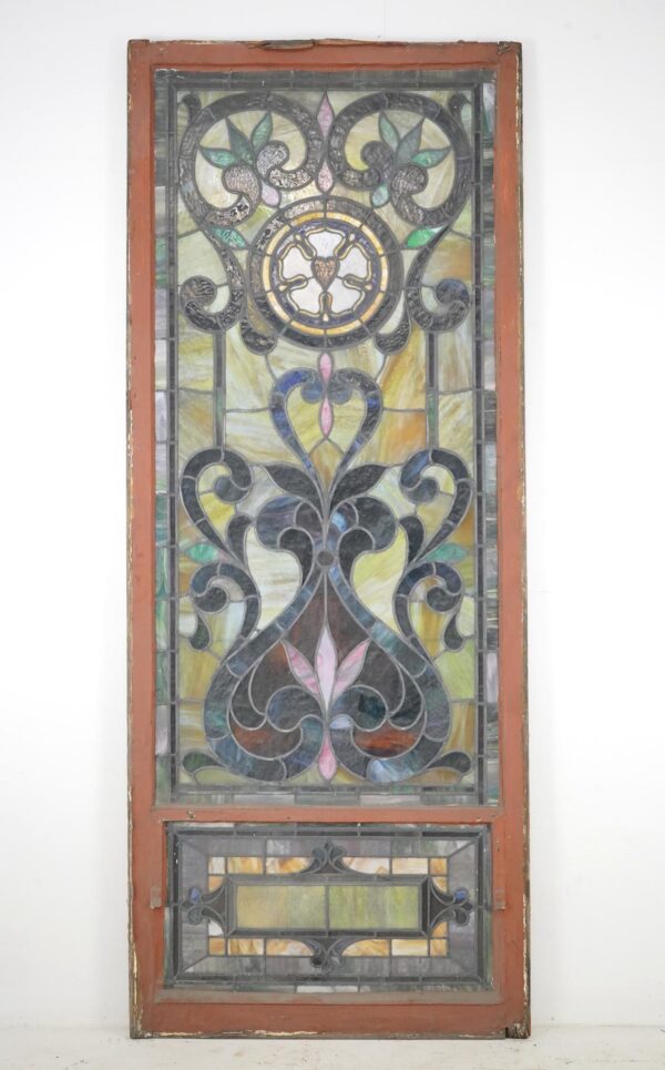 Rectangular leaded stained glass church window with a terra cotta painted wood frame and multicolored panels, shown full length with decorative scrollwork and a smaller framed section below.