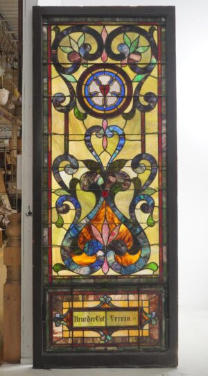 Rectangular leaded stained glass church window with ornate multicolor panels and a lower inscription panel, shown in a dark wood frame.