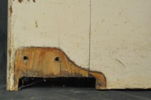 Close-up of a vintage swinging wood door showing the lower edge, with painted white and dark wood tones, surface wear, and a recessed cutout at the bottom.