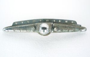 Vintage Studebaker trunk handle with a wing-shaped form, a central chrome button, and a broken end piece, shown in fair condition with heavy surface wear.