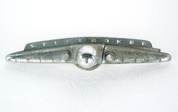 Vintage Studebaker trunk handle with a wing-shaped form, a central chrome button, and a broken end piece, shown in fair condition with heavy surface wear.
