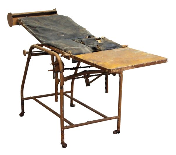 Antique wheeled medical examination chair with a steel frame, worn black padding, and an angled seat and leg rest.