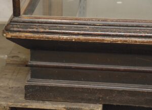 Close-up of an antique retail showcase with a clear glass front, dark wood frame, and heavily worn beveled base.