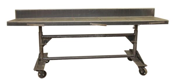 Industrial steel rolling desk with a tall backsplash, adjustable height legs, front safety catches, and four casters.