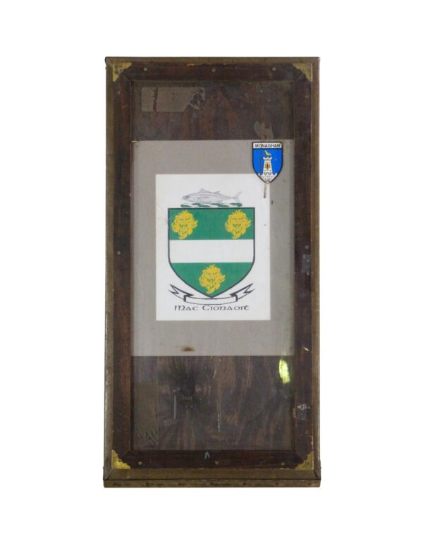 Vintage poster billboard frame with Mac Cionaoit coat of arms and a County Monaghan crest visible behind glass.