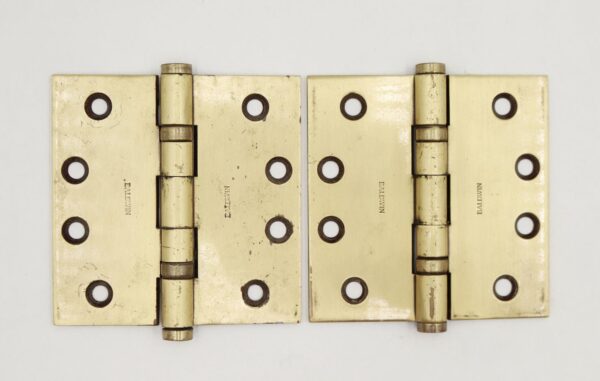 Pair of brass door hinges shown side by side, each with five knuckles, flat tips, and a template hole pattern, with surface wear visible on the metal.