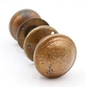 Antique brass passage door knob set shown in a close-up, with four round pieces stacked in a row and one knob face featuring an engraved floral pattern.