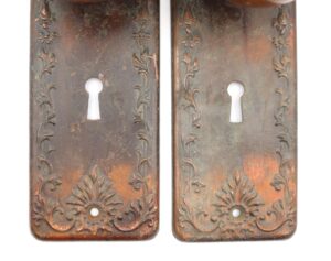 Two antique brass back plates with keyhole cutouts and floral embossing are shown side by side, each with a central dummy spindle hole and visible surface wear.