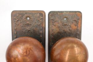 Two antique brass dummy door knobs with ornate steel back plates shown in close-up, with surface wear visible on both pieces.
