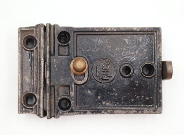 Antique cast iron surface-mount door rim lock with a brass latch and visible mounting holes, shown in close-up with surface wear and the lock switch detail.