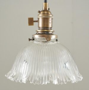 Close-up of an industrial pendant light with an antique brass fitter, a black cord, and a clear ribbed Holophane-style glass shade.