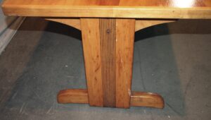 Wooden coffee table shown from the trestle base, with a medium wood tone, vertical ribbed detail at the center, and visible surface wear.