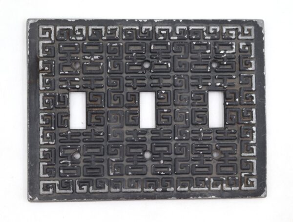 Black Greek key three-gang light switch plate with three rectangular openings and four screw holes, shown front on with heavy surface wear.