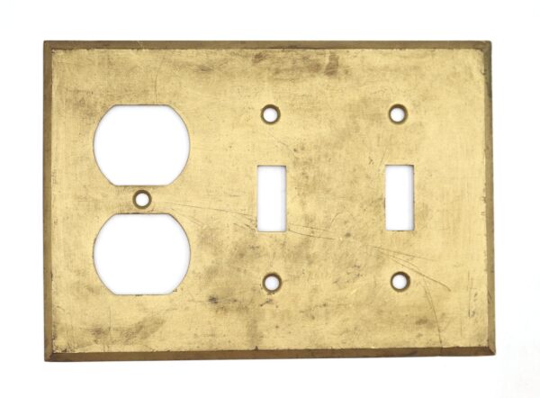 Vintage brass switch plate with one duplex outlet opening, two narrow switch cutouts, and four screw holes, shown with heavy surface wear.